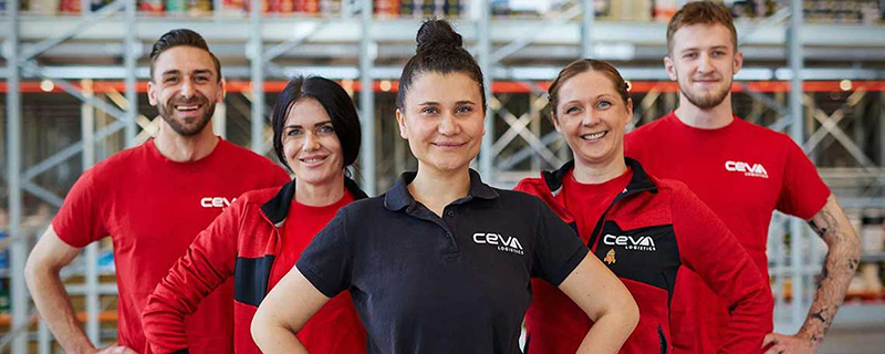 Fünf Mitarbeitende von CEVA Logistics stehen lächelnd in einer Lagerhalle und blicken in die Kamera