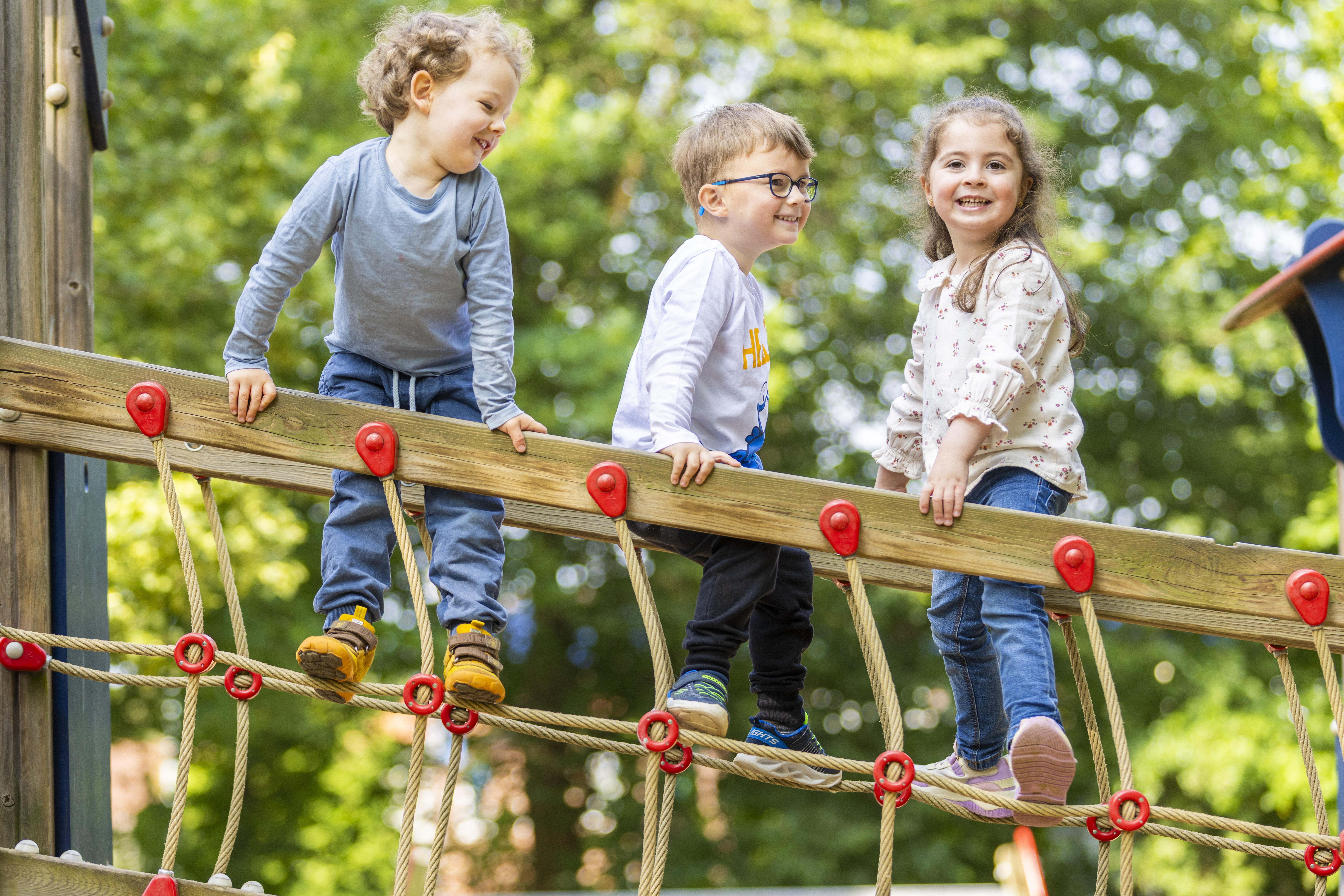 Drei Kinder balancieren auf einem Holz- und Seilklettergerüst in einem grünen Park